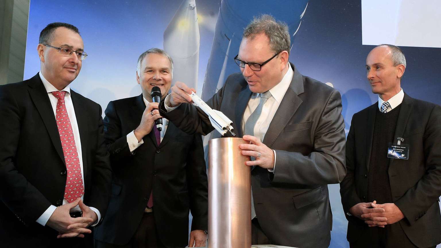 Vier Männer stehen zusammen; einer fügt das Modell einer Rakete in einen Zylinder ein
