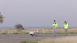 Zwei Männer auf einer Betonpiste mit einem unbemannten Flugobjekt; im Hintergrund die Ostsee